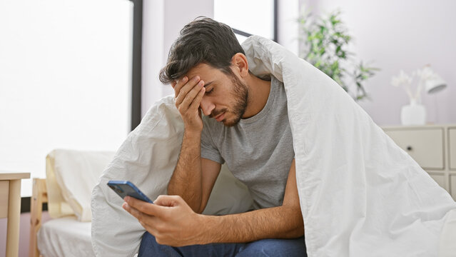 A young hispanic man with a beard looks distraught while sitting on a bed, holding a smartphone in a home interior.