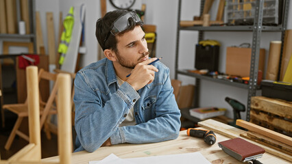 Young bearded man in denim thoughtfully planning at woodworking workshop table with tools.