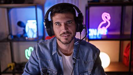 Portrait of a young hispanic man with a beard wearing headphones in a colorful gaming room at night.