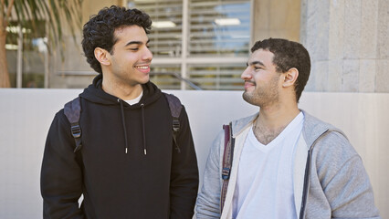 Two smiling hispanic men enjoying friendship outdoors in a sunny urban setting, exuding casual...