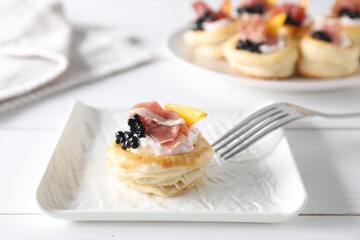 Delicious puff pastry with cream cheese, ham, black caviar, peach and fork on white wooden table, closeup