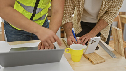 Two men consulting over a blueprint in a carpentry workshop, with digital tablet and woodworking tools present.