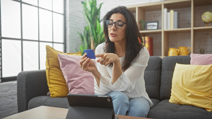 A mature hispanic woman examines a credit card while sitting indoors on a sofa in a cozy living...