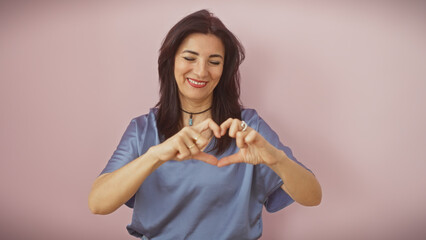 A smiling middle-aged hispanic woman makes a heart sign with her hands against a pink background.