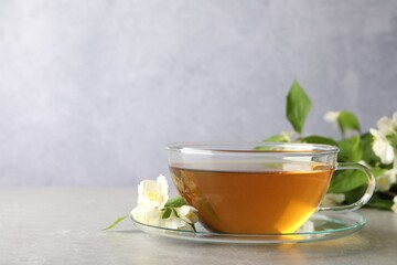 Aromatic jasmine tea in cup, flowers and green leaves on light grey table, space for text