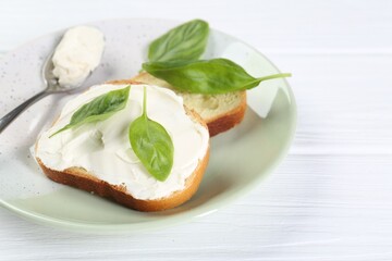 Pieces of bread with cream cheese and basil leaves on white wooden table. Space for text