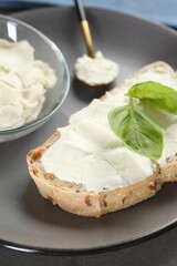 Piece of bread with cream cheese and basil leaves on table, closeup