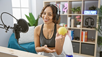 A smiling young woman with headphones holding an apple in a modern radio studio, representing...