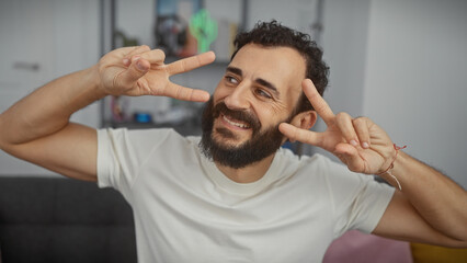 A cheerful man with a beard making peace signs with his hands indoors, expressing positivity and...