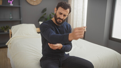 Middle-aged bearded man experiencing arm pain while sitting on the bed in a bedroom interior