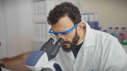 Middle-aged bearded man in lab coat and safety glasses examining specimen through microscope in...