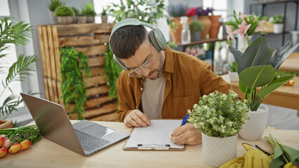 A young man with a beard and headphones writes intently in a flower-filled indoor environment, reflecting a creative or entrepreneurial spirit.