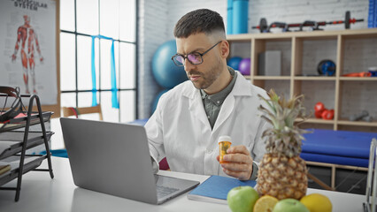 Hispanic man with beard in lab coat examines medication at laptop in physiotherapy clinic.