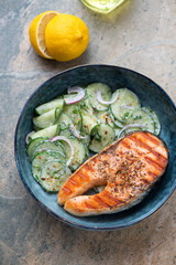 Dark-blue bowl with cucumber salad and grilled salmon steak, vertical shot on a grey and beige granite surface, elevated view
