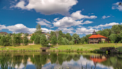 Fototapeta premium View of the pond, blooming meadow, forest and cottages in village at summer timelapse hyperlapse