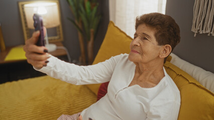 An elderly hispanic woman with short hair takes a selfie while lying on a bed in a cozy bedroom with yellow cushions and a nightstand in the background.