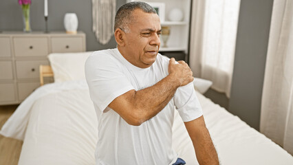 Mature hispanic man sitting on a bed in pain clutching his shoulder indoors at home