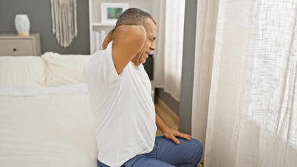 Mature hispanic man in pain sitting on bed at home, rubbing neck in discomfort, depicting healthcare issues.