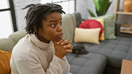Pensive african woman with dreadlocks sitting in a modern living room on a couch, gazing...