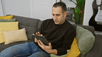 Hispanic man relaxing at home on a couch with tablet, casual, modern interior with guitar.