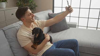 A young hispanic man sits in his living room, holding a dachshund dog and taking a selfie.