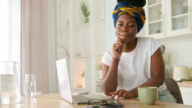 Pensive African American girl sitting at kitchen table with laptop. Young dark skinned woman thinks over work or daily issues, looking through window focused and deep in thoughts