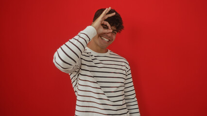 Young hispanic man in a striped shirt on an isolated red background making an okay gesture and smiling.
