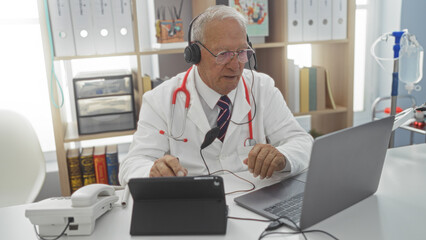 Obraz premium Elderly caucasian man in a doctor's office wearing a stethoscope and headset, attending a video call on a laptop with medical books and equipment in the background.