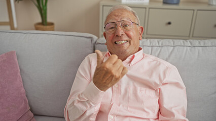 An elderly, grey-haired, caucasian man wearing glasses and a pink shirt sits smiling on a living room sofa, expressing joy through his body language in a cozy home setting.