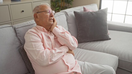 Elderly caucasian man with grey hair sitting on a couch in a living room, looking thoughtful indoors at his home.