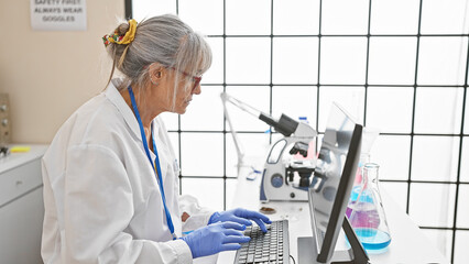 Mature woman scientist working with microscope and computer in laboratory setting.
