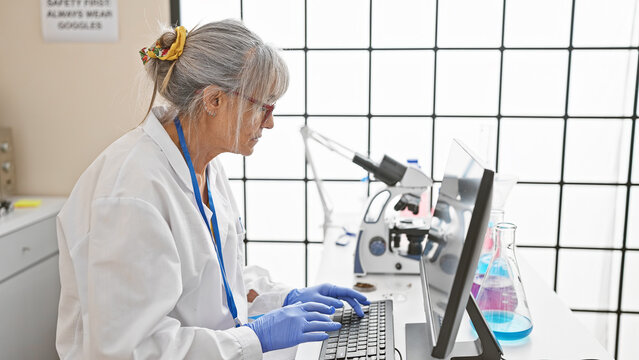 Mature woman scientist working with microscope and computer in laboratory setting.