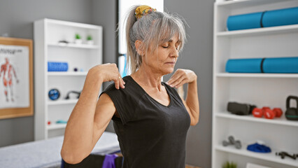 A mature woman with grey hair stretches her neck in a rehab clinic's interior setting, indicating healthcare or physical therapy.