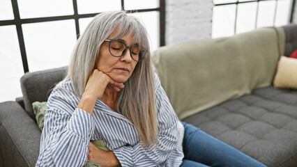 A contemplative mature woman with grey hair and glasses sits on a sofa in a cozy living room.