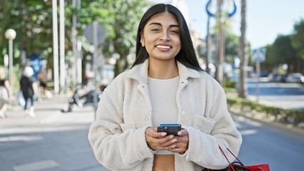 A smiling south asian woman with long hair browses her smartphone on a sunny urban street.