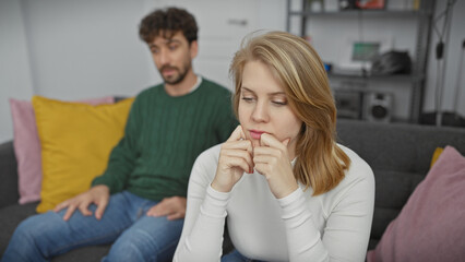 A concerned woman and a contemplative man sit apart on a couch in a modern living room, symbolizing relationship tension.