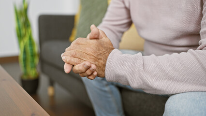 A senior man clasps his hands in a cozy living room, portraying contemplation and tranquility indoors.