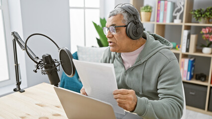 Middle-aged man with headphones reading a script indoors at a podcasting studio setup.