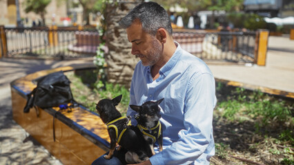 Middle-aged hispanic man sitting outdoors in a city park with two chihuahuas on his lap, enjoying a sunny day.