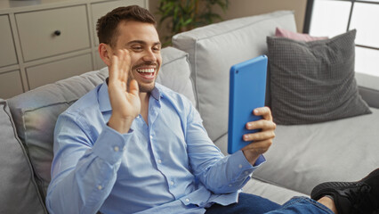 Handsome young hispanic man waving while video chatting on a tablet in a cozy living room setting.