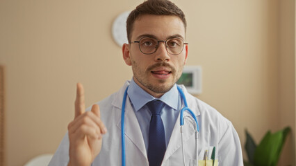 Handsome hispanic man with a beard, wearing a white lab coat and stethoscope, gestures while standing in a clinic lobby.