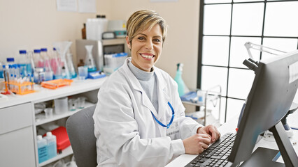 Smiling hispanic woman with short blonde hair working in white lab coat at indoor laboratory...