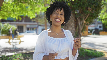 A beautiful young african-american woman with curly hair smiles brightly while standing outdoors in an urban park.