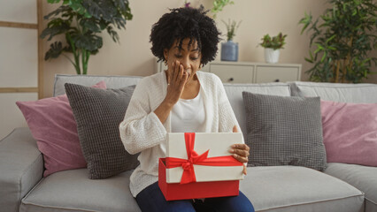 Young african american woman with curly hair surprised by a gift in a box with a red ribbon,...