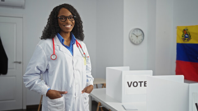A young african american woman with curly hair, in a medical coat with a stethoscope, stands in an electoral room with venezuelan flags and voting booths.