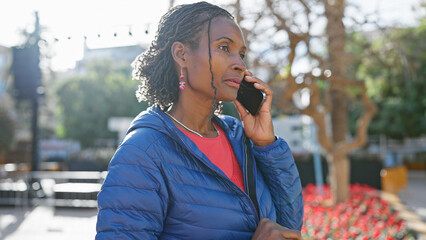 Mature african woman in a blue jacket talking on a smartphone outdoors in a sunny urban setting.