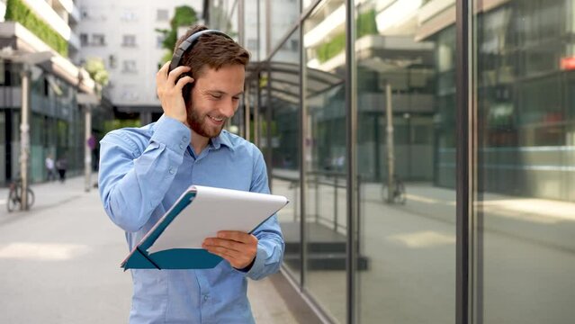 A young professional man stands confidently outside a corporate building, holding a clipboard and wearing headphones.