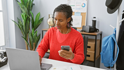 Professional african american woman using smartphone and laptop in modern office setting