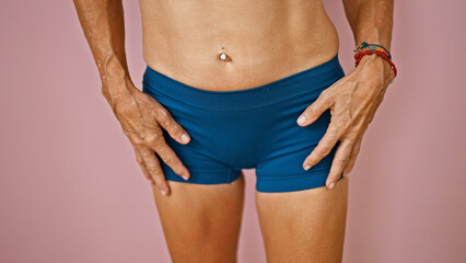 A close-up portrait of an adult woman's midsection against a pink wall, highlighting her blue shorts and a bracelet.