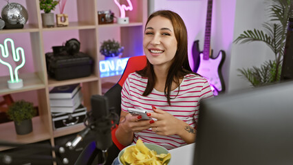 A smiling young woman using smartphone in her colorful gaming room filled with neon lights, guitar,...